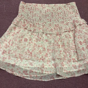 Blommig kjol från Gina Tricot - Söt blommig kjol från Gina Tricot i rosa och vit med volangdetaljer. Kjolen har en elastisk midja för en bekväm passform. Perfekt för varma dagar och ger en romantisk touch till din outfit.