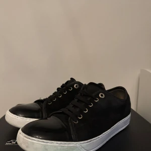 Svarta sneakers från Lanvin - Snygga svarta sneakers från Lanvin med glänsande tåhätta och vit sula. Skorna har snörning och en stilren design som passar perfekt för en vardag skorna är i 6-10 skick för det är lite slitet under men annars ser dom nya ut, jag tycker det är ett bra kap för det priset så hör av dig om du har några frågor🤝