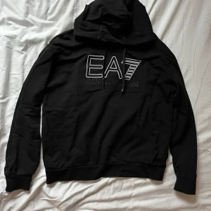 Svart hoodie från Emporio Armani - Snygg svart hoodie från Emporio Armani med EA7-logga på bröstet. Perfekt för en stilren och avslappnad look. Den har en klassisk huva med dragsko och långa ärmar. Nypris 899:- SEK 