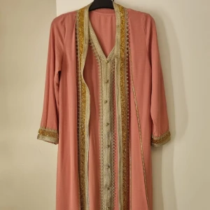 Rosa kaftan med gulddetaljer - Elegant rosa kaftan med guldfärgade broderade detaljer längs kanterna och ärmsluten. Klänningen har en v-ringad design och inkluderar en matchande ärmlös underklänning. Perfekt för speciella tillfällen.
