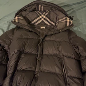 Svart dunjacka från Burberry - Kvitto ingår. Säljer en snygg svart dunjacka från Burberry med klassiskt rutigt mönster i fodret. Jackan har en huva och stängs med dragkedja och knappar. Perfekt för kyliga dagar! 