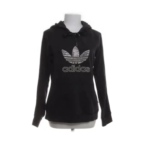 Adidas Svart hoodie strl 36/38 - Säljer en svart hoodie från Adidas med det klassiska logotyptrycket på framsidan. Hoodien har en justerbar huva med dragsko och långa ärmar. Materialet är en blandning av bomull och polyester för en bekväm passform.