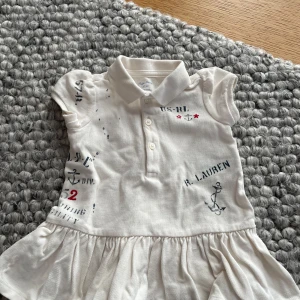 Vit klänning från Ralph Lauren - Söt vit klänning från Ralph Lauren med korta ärmar och krage. Klänningen har tryck med ankare och textdetaljer i blått och rött. Perfekt för små äventyrare! 👶✨