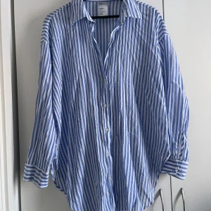 Blå och vit randig skjorta - Säljer en stilren blå och vit randig skjorta med knappar framtill. Skjortan har en avslappnad passform och långa ärmar, perfekt för en casual look. Tillverkad i ett lätt och bekvämt material.