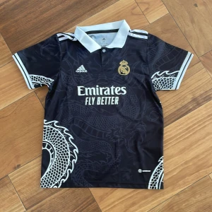 Svart Real Madrid fotbollströja- special 2022-2023 - Snygg svart Real Madrid fotbollströja från Adidas med ett unikt drakmönster i grått. Tröjan har korta ärmar och en vit krage. Perfekt för alla fotbollsfans som vill sticka ut på planen eller läktaren.