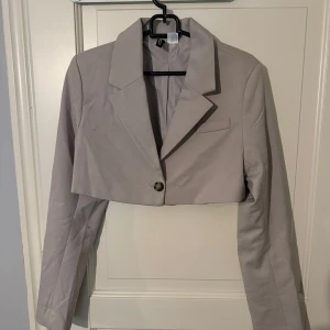 Beige kavaj med knapp - Kroppad beige kavaj med en knapp framtill. Den har en klassisk krage och långa ärmar. Perfekt för en stilren look.