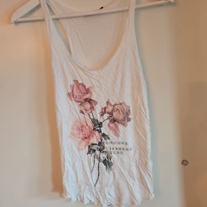 Vit linne med blommotiv - Säljer ett vitt linne med ett vackert blommotiv. Texten 'Live in the past, present, future' är tryckt under blommorna. Perfekt för varma sommardagar.