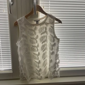Vit ärmlös topp från H&M - Säljer en vit ärmlös topp från H&M med broderat blommönster. Toppen är tillverkad i ett lätt och luftigt material. Aldrig använd🤍