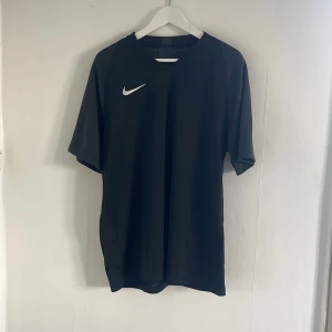 Svart fotbollströja från Nike - Säljer en stilren svart fotbollströja från Nike med korta ärmar. Tröjan är tillverkad i ett lätt och andningsbart material, perfekt för träning eller match. Den har en vit Nike-logga på bröstet och är gjord av polyester för optimal komfort.