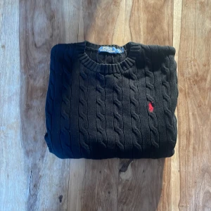 Svart kabel stickad tröja  Polo Ralph Lauren(nästan aldrig använd) - TRÖJAN FINNS ÄVEN HOS @Arvid Carlsson MED FRI FRAKT! Säljer en stilren svart stickad tröja från Polo Ralph Lauren med kabelstickat mönster. Tröjan har en klassisk rund halsringning och ett rött broderat logotypmärke på bröstet. Perfekt för en snygg och avslappnad look. Pris är ej hugget i sten!