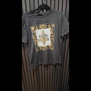 Svart t-shirt med guldtryck - Snygg svart t-shirt med ett iögonfallande guldtryck på framsidan. Perfekt för att ge din outfit en lyxig touch. Kortärmad och tillverkad i ett mjukt material för bekväm passform.
