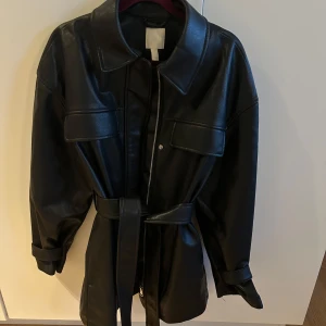Svart skinnjacka med bälte -  Svart skinnjacka med dragkedja och bälte i midjan. Jackan har långa ärmar och stora fickor framtill. .