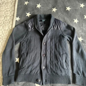 Marinblå cardigan från French Connection - Snygg grå cardigan från French Connection med knappar framtill och quiltade detaljer på axlarna. Den har en ribbad krage och muddar, vilket ger en stilren look. Perfekt för kyligare dagar. Man kan både använda den som en tröja och en cardigan/tunn jacka under våren. Skicket är bra och tröj är använd. Ifall du har fler frågor är det bara att höra av dig!!!!