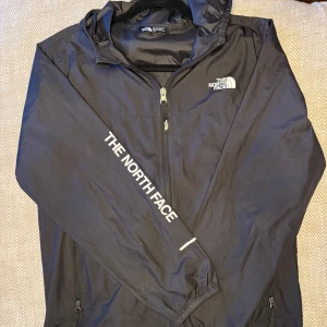 The North Face jacka - Säljer en svart vindjacka från The North Face med dragkedja och huva. Jackan har långa ärmar och logotypen tryckt på bröstet och längs ärmen. Perfekt för blåsiga dagar.