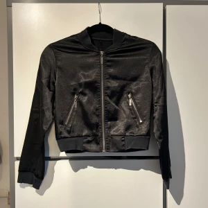 Svart bomberjacka med dragkedjor - Snygg svart bomberjacka med glansig finish och dragkedjor framtill. Jackan har en klassisk bomberstil med ribbade muddar vid ärmslut och nederkant.