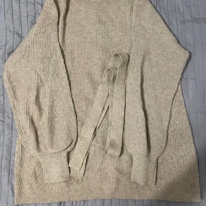 Beige stickad tröja från VILA - Säljer en stilren beige stickad tröja från VILA. Tröjan har långa ärmar och ett ribbat mönster. Den kommer med ett matchande bälte för en snygg passform. Perfekt för kyligare dagar.