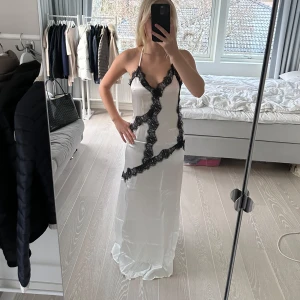 Vit långklänning med spetsdetaljer från NA-KD - Elegant vit långklänning från NA-KD med svarta spetsdetaljer som löper diagonalt över klänningen. Den har smala axelband och en figurnära passform som ger en stilren look. Den är från deras ART Collection vilket är en mer exklusiv kollektion. Nypris är 1299kr. Den är aldrig använd! Helt slutsåld
