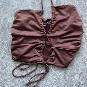 Brun halterneck topp från Bik Bok - Snygg brun omlott topp från Bik Bok med smala axelband och knytning framtill. Perfekt för en stilren look. Toppen har en åtsittande passform och är tillverkad i ett mjukt material. Aldrig använt. 