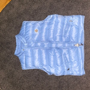 Ljusblå dunväst från Moncler - Snygg ljusblå dunväst från Moncler med dragkedja och två praktiska fickor framtill. Västen har en klassisk design med Moncler-logga på bröstet. Perfekt väst nu inför våren för mer detaljerde bilder vänligen skicka ett meddelande.  skickas med plick säkra köp för båda parets säkerhet vid snabbäffar kan jag gå ner till 1100 kr / Mvh Thom 