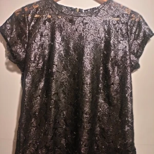 Svart spetsblus från Gina Tricot - Snygg svart glittrig spetsblus från Gina Tricot i storlek M. Blusen har korta ärmar och en dragkedja i ryggen. Perfekt för en elegant look med sitt vackra spetsmönster.