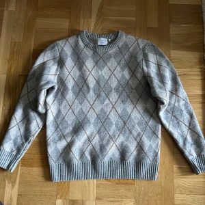 Grå rutig tröja från Bläck - Säljer en stilren grå tröja från Bläck med ett klassiskt rutmönster i brunt och ljusgrått. Tröjan har långa ärmar och ribbade muddar vid ärmslut och nederkant. Perfekt för kyligare dagar.