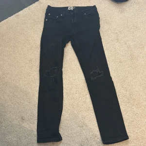 Svarta jeans med slitningar - Snygga svarta jeans med slitningar på knäna. De har en dragkedja och knapp framtill samt klassiska femficksdesign. Perfekta för en avslappnad stil med en edgy touch.