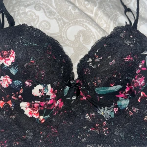 Blommig spetskorsett från H&M - Säljer en vacker svart spetskorsett från H&M med blommigt mönster i rosa och gröna nyanser. Den har justerbara axelband och hakknäppning bak. Perfekt för att lägga till en romantisk touch till din outfit.