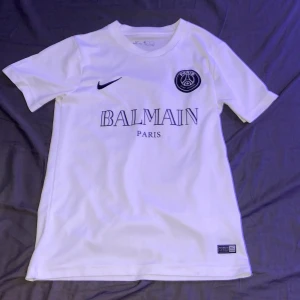 Psg, Balmain tröja - Snygg vit fotbollströja från Nike i samarbete med Balmain. Tröjan har korta ärmar och är tillverkad i Dri-FIT-material för optimal komfort. Den har Balmain Paris-logga och PSG-emblem på bröstet. Perfekt för både träning och vardag.
