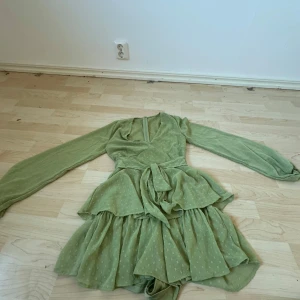 Grön klänning med volanger - Charmig grön klänning med volanger och knytband i midjan. Den har långa ärmar och ett diskret prickigt mönster. Perfekt för en somrig look!