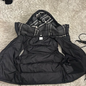 Burberry Beat Check Jacka/Väst - Snygg svart dunväst med avtagbar huva och knappar framtill. Insidan har ett stilrent rutigt mönster i grått och vitt. Perfekt för kyliga dagar när du vill hålla värmen utan att kompromissa med stilen.. Armarna är använda nån gång och tillkommer med västen