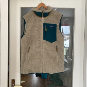 Beige fleeceväst från Patagonia - Säljer en supermysig beige fleeceväst från Patagonia i storlek M. Den har en dragkedja fram och en snygg blå ficka med dragkedja på bröstet. Perfekt för höst och vår när du vill hålla dig varm och bekväm. Västen är helt ny bara testat några gånger 