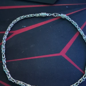 Silverfärgat halsband med kedjedesign - Elegant silverfärgat halsband med en intrikat kedjedesign. Halsbandet har en säker karbinhake som lås, vilket gör det både stilrent och praktiskt. Perfekt för att ge en sofistikerad touch till din outfit.