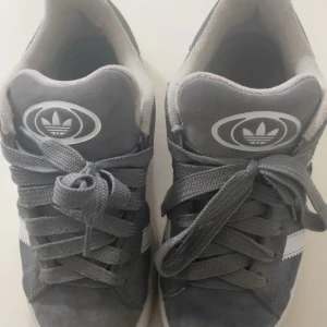 Grå sneakers från (Adidas Campus) - Snygga grå sneakers från Adidas med klassisk design och vit sula. Skorna har snörning och det ikoniska Adidas-märket syns både på plösen och hälen. Perfekta för en avslappnad stil.