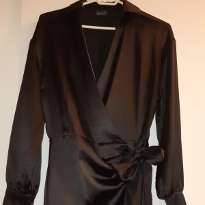 Svart satin omlottklänning Gina Tricot - Elegant svart omlottklänning med långa ärmar och knytning i midjan. Klänningen har en v-ringad design och är tillverkad i ett glansigt material som ger en lyxig känsla. Perfekt för en stilren look.