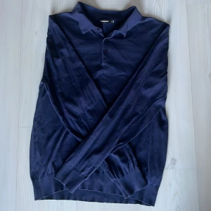 Mörkblå pikétröja från J.Lindeberg - Säljer en stilren mörkblå pikétröja från J.Lindeberg i bomull och silke. Tröjan har långa ärmar och en klassisk krage med knappar framtill. Perfekt för en avslappnad men ändå elegant look. Skriver L för den psssr L