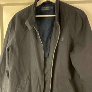 Blå jacka från Polo Ralph Lauren - Snygg blå jacka från Polo Ralph Lauren med dragkedja och knappar vid kragen. Perfekt för en stilren look. Jackan har en klassisk design med diskret logga på bröstet.