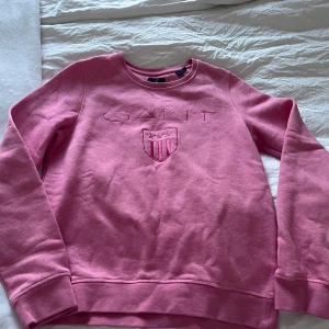Rosa sweatshirt från GANT - Säljer en snygg rosa sweatshirt från GANT med broderad logga och emblem på bröstet. Tröjan har långa ärmar och en rund halsringning. Perfekt för en avslappnad stil.