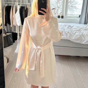 Champagnefärgad klänning från Dry Lake - Elegant champagnefärgad klänning från Dry Lake med långärmad design och knytband i midjan. Klänningen har en vacker öppen rygg och är tillverkad i ett glansigt material som ger en lyxig känsla. Perfekt för speciella tillfällen. Nypris är 1300kr. Den är i perfekt skick!