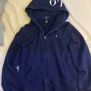 Mörkblå hoodie från Polo Ralph Lauren - Säljer en snygg mörkblå hoodie från Polo Ralph Lauren med dragkedja och huva. Den har en liten logga på bröstet och stor text på huvan. Perfekt för en avslappnad stil.