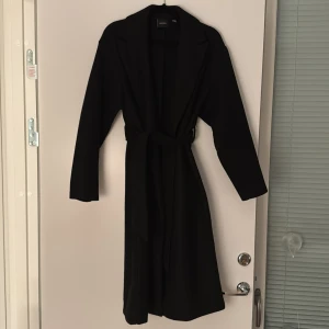 Svart kappa från Vero Moda - Elegant svart kappa från Vero Moda med knytbälte i midjan. Delvis nopprig men som enkelt går att plocka bort eller med apparat. Från Vero Moda i storlek XS. Går under knäna på mig som är 165