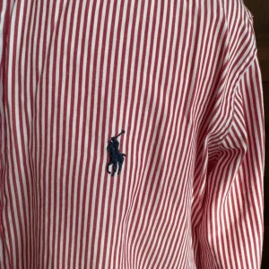 Röd- och vitrandig skjorta från Ralph Lauren - Snygg röd- och vitrandig skjorta från Ralph Lauren med broderad logga på bröstet. Skjortan har långa ärmar och knappar framtill. Perfekt för en stilren look.