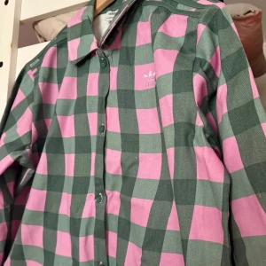 Rutig skjorta från Adidas - Snygg rutig skjorta från Adidas i grönt och rosa. Skjortan har långa ärmar och knappar framtill. Perfekt för en avslappnad stil med en sportig touch.