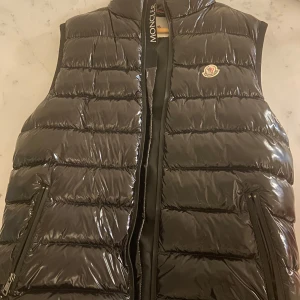  Moncler väst - Snygg svart dunväst från Moncler med glansig yta och dragkedja framtill. Västen har en klassisk design med Moncler-logga på bröstet och praktiska fickor med dragkedja. Perfekt för kyligare dagar.