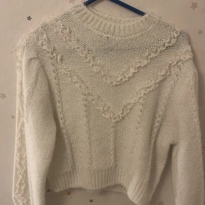 Vit stickad tröja från Divided - Säljer en vit stickad tröja från Divided med dekorativa volangdetaljer på framsidan. Tröjan har en rund hals och långa ärmar, perfekt för kyligare dagar. Den är både stilren och mysig, ett måste i garderoben! 💫