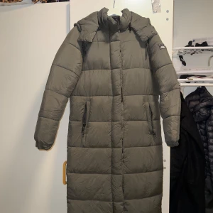 Grön lång dunjacka från superdry - Säljer en lång och varm grön dunjacka från superdry . Jackan har en hög krage och dragkedja framtill. Perfekt för kalla dagar med sin pufferdesign och långa ärmar.