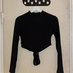 Svart ribbad topp från Shein - Säljer en svart ribbad topp från Shein med långa ärmar, det blir ett kors på magen med banden nertill. Perfekt för en stilren look. 🖤