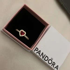 Guldfärgad ring med hjärta från Pandora - Elegant guldfärgad ring från Pandora med ett rött hjärta i mitten, omgiven av gnistrande stenar. Perfekt för att ge en romantisk touch till din stil. Levereras i originalförpackning.