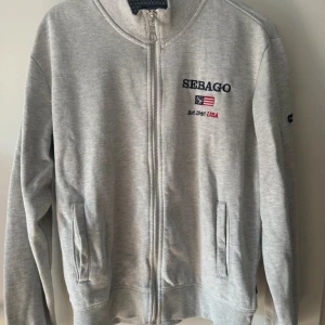 Grå zip-tröja från Sebago - Säljer en stilren grå zip-tröja från Sebago med broderad logga på bröstet och ärmen. Tröjan har långa ärmar och ribbade muddar vid ärmslut och nederkant. Perfekt för en avslappnad look.