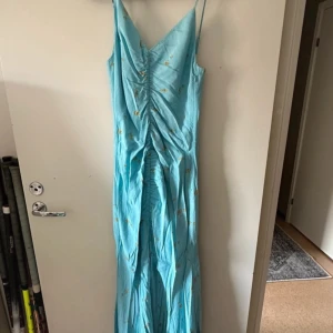 Ljusblå långklänning från ADOORE - Snygg ljusblå långklänning från ADOOREi storlek 36 med smala axelband och ett subtilt blommönster. Klänningen har en v-ringad design och rynkade detaljer framtill som ger en smickrande passform. Oanvänd utan prislapp.