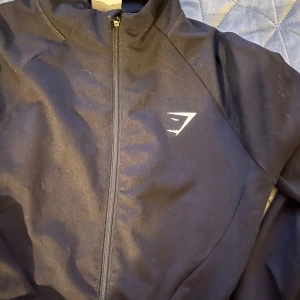 Svart träningskofta från Gymshark - Snygg svart träningskofta från Gymshark med dragkedja framtill. Perfekt för gymmet eller löprundan. Jackan har en stilren design med Gymshark-logga på bröstet.